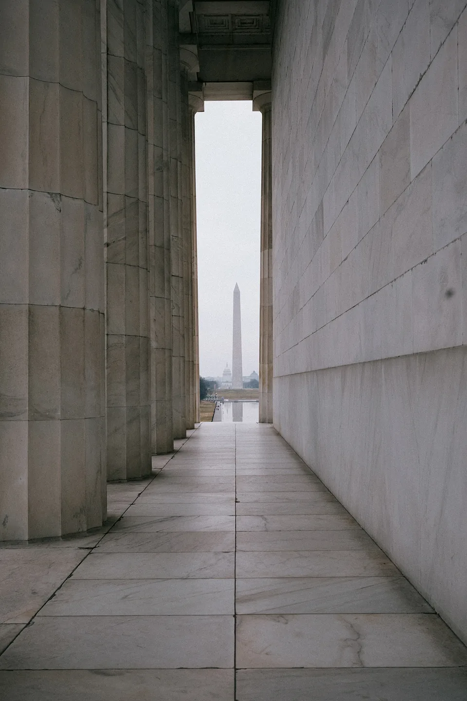 A narrow view between stone columns and a marble wall frames the Washington Monument in the distance.