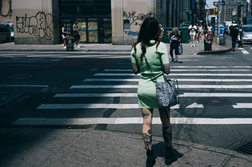 A person in a pale green dress stands at a sunlit crosswalk, with deep shadows across the road.