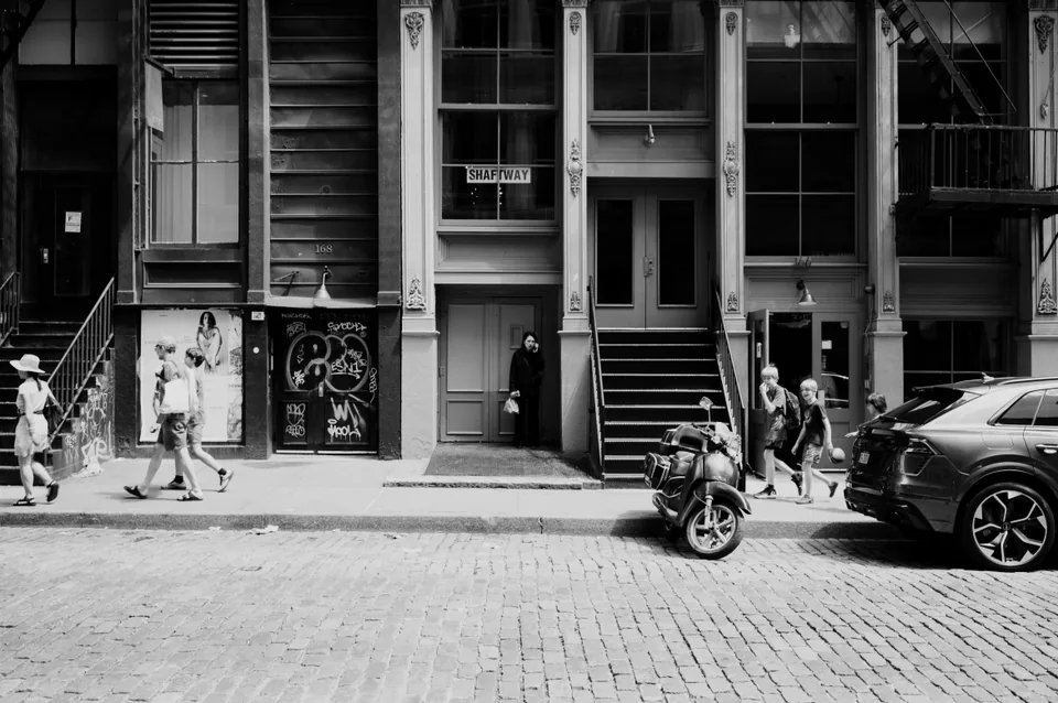 Black-and-white street scene with a person in a doorway, pedestrians, and a parked scooter below tall windows.