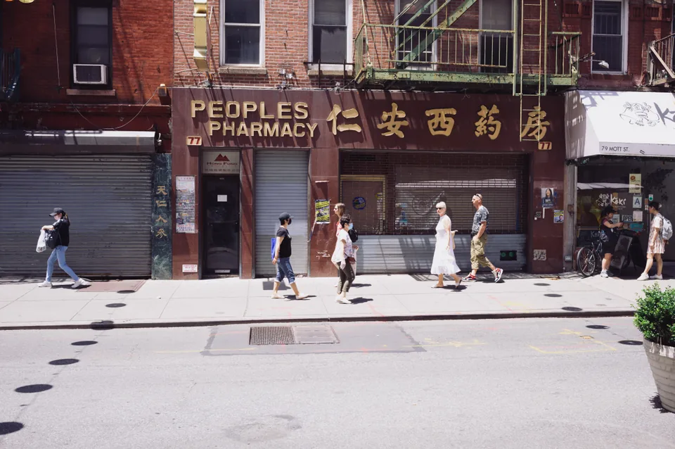 Pedestrians pass the shuttered Peoples Pharmacy beneath brick apartments and fire escapes.