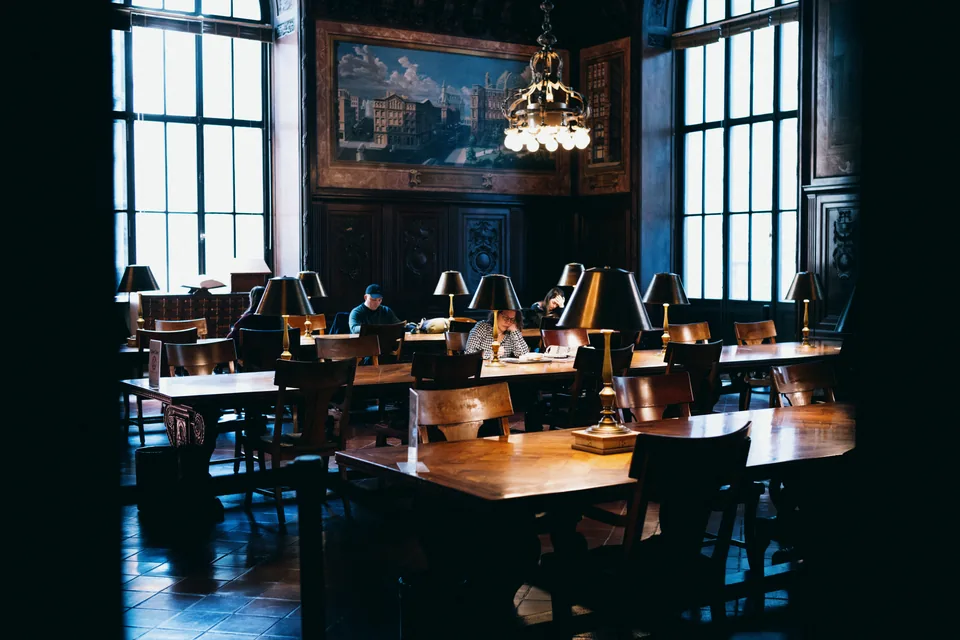 Readers sit at wooden tables under brass lamps in a dark paneled room lit by tall windows.