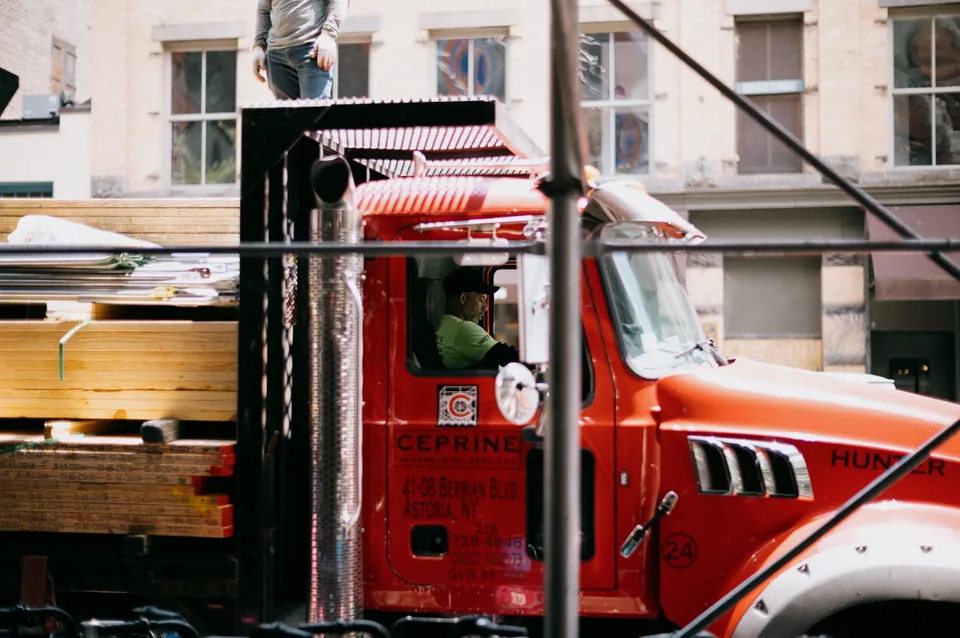 A red flatbed truck carrying lumber, framed by dark scaffolding poles.